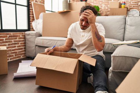 Photo for Young hispanic man unboxing package with worry expression at new home - Royalty Free Image