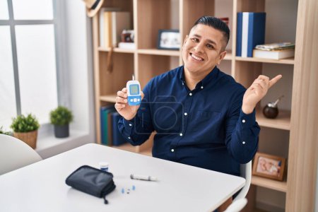 Foto de Hispanic young man holding glucometer device smiling happy pointing with hand and finger to the side - Imagen libre de derechos