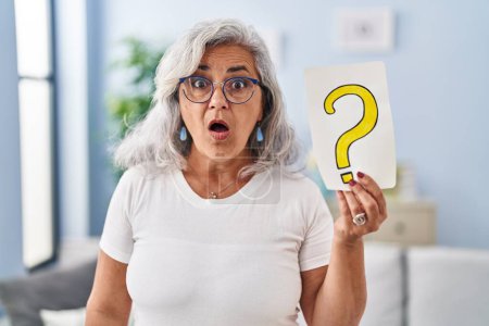 Foto de Middle age woman with grey hair holding question mark scared and amazed with open mouth for surprise, disbelief face - Imagen libre de derechos