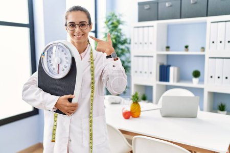 Photo for Young hispanic woman as nutritionist doctor holding weighing machine pointing finger to one self smiling happy and proud - Royalty Free Image
