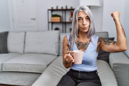 Téléchargez les photos : Femme aux cheveux gris d'âge moyen buvant du café assis sur le canapé à la maison personne forte montrant muscle du bras, confiante et fière de la puissance - en image libre de droit