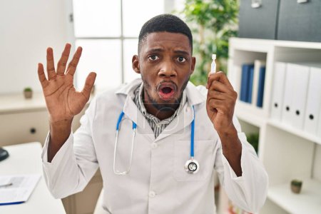 Téléchargez les photos : African american doctor man holding eye drops scared and amazed with open mouth for surprise, disbelief face - en image libre de droit
