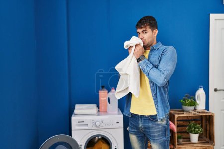Téléchargez les photos : Young hispanic man smelling dirty towel standing at laundry room - en image libre de droit