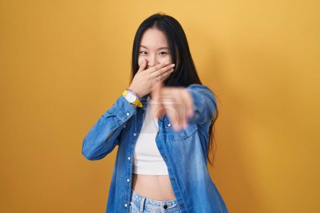 Téléchargez les photos : Jeune femme asiatique debout sur fond jaune se moquant de vous, pointant du doigt la caméra avec la main sur la bouche, expression de honte - en image libre de droit