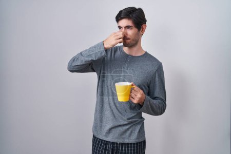 Foto de Joven hispano con pijama bebiendo una taza de café oliendo algo apestoso y asqueroso, olor intolerable, conteniendo la respiración con los dedos en la nariz. mal olor - Imagen libre de derechos