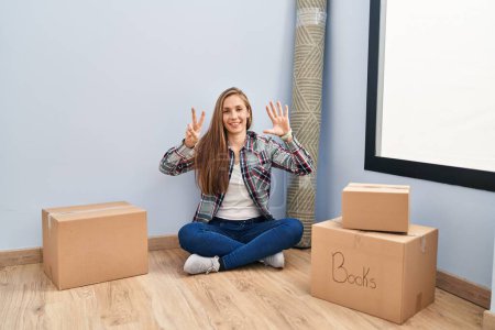 Foto de Young blonde woman sitting on the floor moving to a new home showing and pointing up with fingers number seven while smiling confident and happy. - Imagen libre de derechos