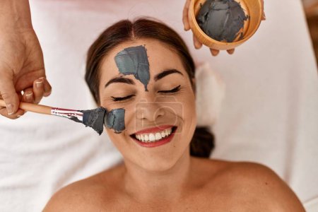 Photo for Young beautiful hispanic woman lying on table having facial treatment at beauty salon - Royalty Free Image