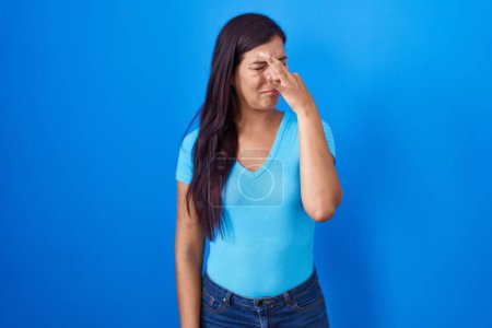 Téléchargez les photos : Jeune femme hispanique debout sur fond bleu sentant quelque chose de puant et dégoûtant, odeur intolérable, retenant souffle avec les doigts sur le nez. mauvaise odeur - en image libre de droit