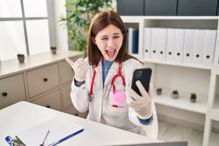 Photo for Young doctor woman doing video call with smartphone pointing thumb up to the side smiling happy with open mouth - Royalty Free Image