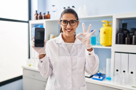 Foto de Young hispanic woman working at scientist laboratory with smartphone doing ok sign with fingers, smiling friendly gesturing excellent symbol - Imagen libre de derechos