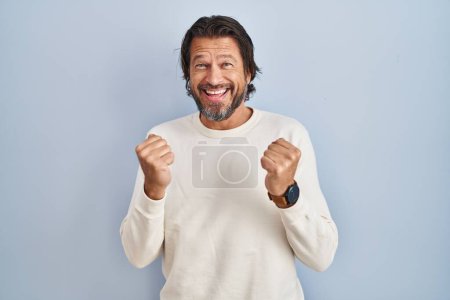 Photo for Handsome middle age man wearing casual sweater over blue background celebrating surprised and amazed for success with arms raised and open eyes. winner concept. - Royalty Free Image