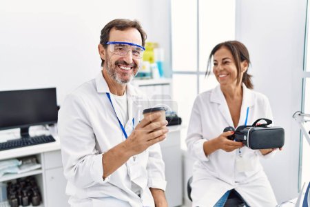 Téléchargez les photos : Hommes et femmes d'âge moyen portant un uniforme scientifique buvant du café et tenant des lunettes de protection au laboratoire - en image libre de droit
