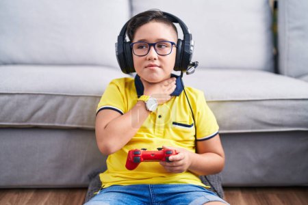 Téléchargez les photos : Jeune enfant hispanique jouant contrôleur de tenue de jeu vidéo portant des écouteurs touchant le cou douloureux, mal de gorge pour la grippe, la motte et l'infection - en image libre de droit