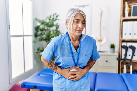 Téléchargez les photos : Middle age grey-haired woman wearing physiotherapist uniform at medical clinic with hand on stomach because nausea, painful disease feeling unwell. ache concept. - en image libre de droit