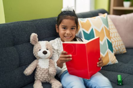 Foto de Adorable hispanic girl reading book sitting on sofa at home - Imagen libre de derechos