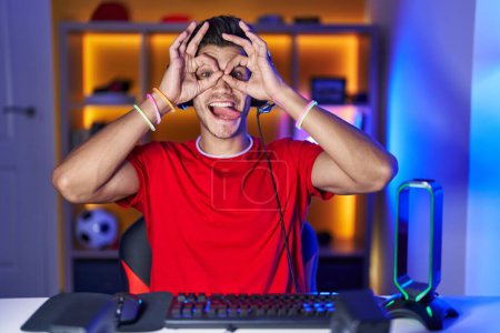 Foto de Joven hispano jugando videojuegos haciendo un buen gesto como binoculares sacando la lengua, ojos mirando a través de los dedos. expresión loca. - Imagen libre de derechos