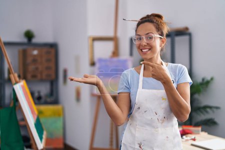 Foto de Brunette woman painting at art studio amazed and smiling to the camera while presenting with hand and pointing with finger. - Imagen libre de derechos