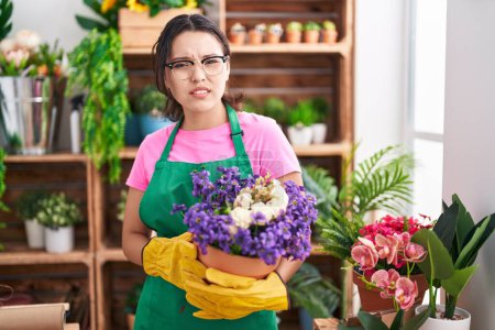Foto de Mujer joven hispana que trabaja en floristería sosteniendo la planta sin pistas y expresión confusa. concepto de duda. - Imagen libre de derechos