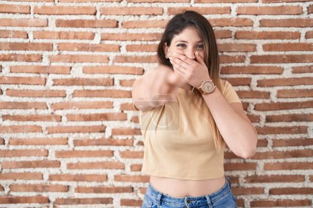 Téléchargez les photos : Jeune femme brune debout sur un mur de briques se moquant de vous, pointant du doigt la caméra avec la main sur la bouche, expression de honte - en image libre de droit