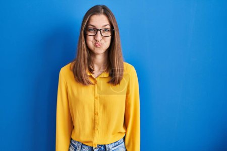 Téléchargez les photos : Young woman wearing glasses standing over blue background puffing cheeks with funny face. mouth inflated with air, crazy expression. - en image libre de droit