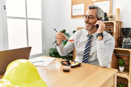 Téléchargez les photos : Moyen Age homme aux cheveux gris architecte parler sur smartphone manger des sushis au bureau - en image libre de droit