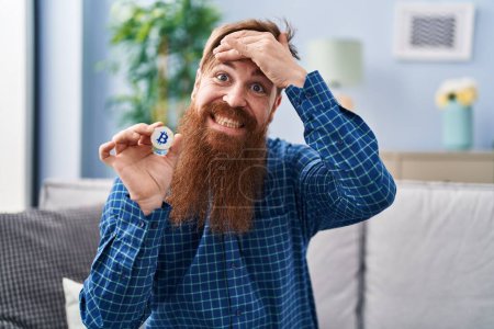 Foto de Caucasian man with long beard holding virtual currency bitcoin stressed and frustrated with hand on head, surprised and angry face - Imagen libre de derechos
