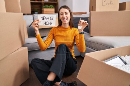 Photo for Young caucasian woman sitting on the floor at new home holding new home banner sticking tongue out happy with funny expression. - Royalty Free Image
