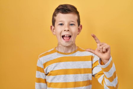 Foto de Young caucasian kid standing over yellow background smiling and confident gesturing with hand doing small size sign with fingers looking and the camera. measure concept. - Imagen libre de derechos