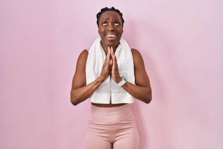 Téléchargez les photos : Belle femme noire portant des vêtements de sport et une serviette sur fond rose mendiant et priant avec les mains ainsi que l'expression d'espoir sur le visage très émotionnel et inquiet. mendier. - en image libre de droit