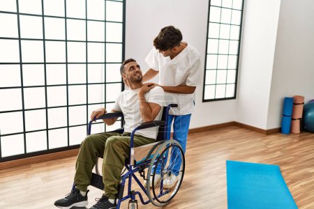 Foto de Dos hombres hispanos fisioterapeuta y paciente sentado en silla de ruedas teniendo una sesión de rehabilitación en la clínica - Imagen libre de derechos