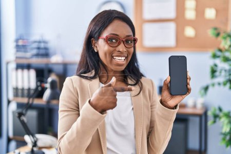 Téléchargez les photos : Africaine jeune femme tenant smartphone montrant écran vierge souriant heureux pointant avec la main et le doigt - en image libre de droit