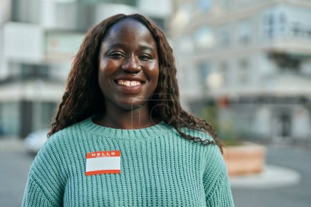 Téléchargez les photos : Jeune femme africaine souriante heureuse portant bonjour je suis autocollant à la ville - en image libre de droit