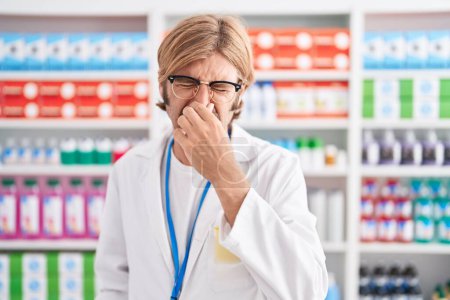 Foto de Caucasian man with mustache working at pharmacy drugstore smelling something stinky and disgusting, intolerable smell, holding breath with fingers on nose. bad smell - Imagen libre de derechos