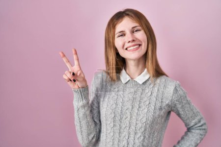 Foto de Beautiful woman standing over pink background smiling looking to the camera showing fingers doing victory sign. number two. - Imagen libre de derechos