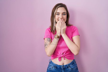Foto de Blonde caucasian woman standing over pink background laughing and embarrassed giggle covering mouth with hands, gossip and scandal concept - Imagen libre de derechos
