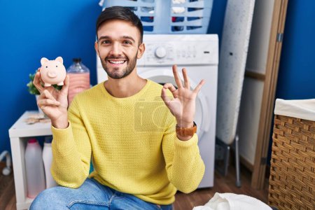 Photo for Hispanic man doing laundry holding piggy bank doing ok sign with fingers, smiling friendly gesturing excellent symbol - Royalty Free Image