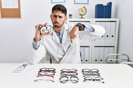 Photo for Young optician man holding optometry glasses pointing down looking sad and upset, indicating direction with fingers, unhappy and depressed. - Royalty Free Image
