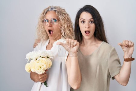 Photo for Mother and daughter holding bouquet of white flowers surprised pointing with hand finger to the side, open mouth amazed expression. - Royalty Free Image