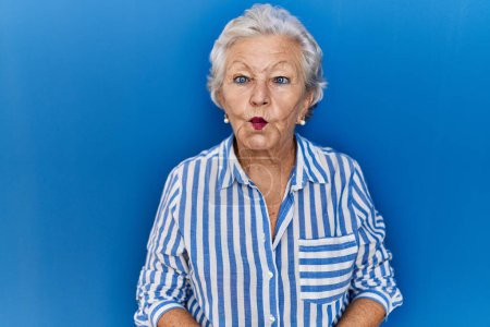 Téléchargez les photos : Femme âgée avec les cheveux gris debout sur fond bleu faisant visage de poisson avec des lèvres, geste fou et comique. expression drôle. - en image libre de droit