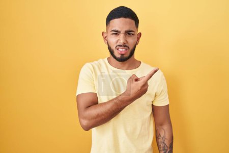 Foto de Young hispanic man standing over yellow background pointing aside worried and nervous with forefinger, concerned and surprised expression - Imagen libre de derechos