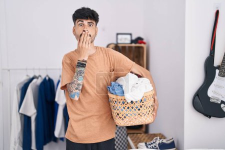 Téléchargez les photos : Young hispanic man with beard holding laundry basket at bedroom covering mouth with hand, shocked and afraid for mistake. surprised expression - en image libre de droit