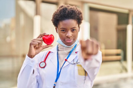 Téléchargez les photos : Femme afro-américaine portant un uniforme de médecin tenant le cœur pointant du doigt vers la caméra et vers vous, geste confiant et sérieux - en image libre de droit
