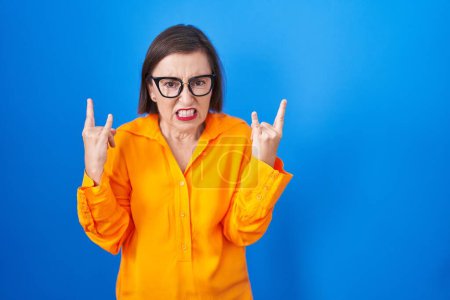 Foto de Middle age hispanic woman wearing glasses standing over blue background shouting with crazy expression doing rock symbol with hands up. music star. heavy concept. - Imagen libre de derechos