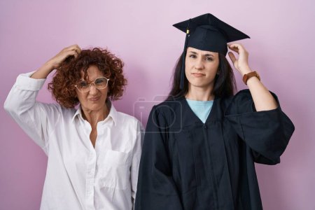 Foto de Hispanic mother and daughter wearing graduation cap and ceremony robe confuse and wonder about question. uncertain with doubt, thinking with hand on head. pensive concept. - Imagen libre de derechos