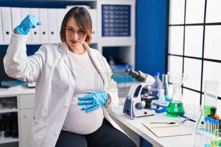 Téléchargez les photos : Femme enceinte travaillant au laboratoire scientifique personne forte montrant muscle du bras, confiant et fier de la puissance - en image libre de droit