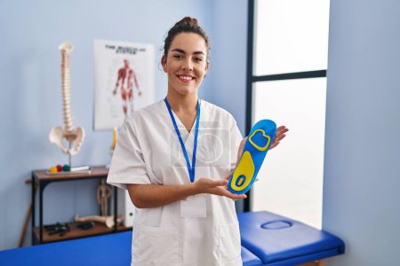 Photo for Young hispanic woman holding shoe insole at physiotherapy clinic smiling with a happy and cool smile on face. showing teeth. - Royalty Free Image