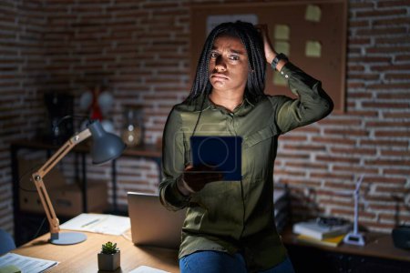 Foto de Young african american with braids working at the office at night confuse and wondering about question. uncertain with doubt, thinking with hand on head. pensive concept. - Imagen libre de derechos