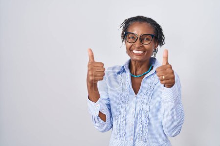 Foto de Mujer africana con rastas de pie sobre fondo blanco con gafas signo de éxito haciendo gesto positivo con la mano, pulgares hacia arriba sonriendo y feliz. expresión alegre y gesto ganador. - Imagen libre de derechos