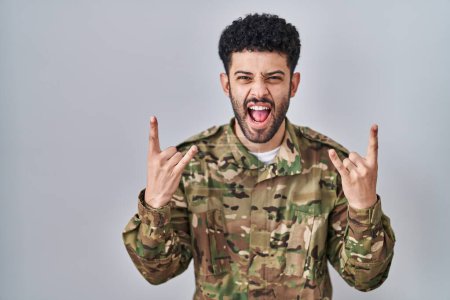 Téléchargez les photos : Arab man wearing camouflage army uniform shouting with crazy expression doing rock symbol with hands up. music star. heavy music concept. - en image libre de droit