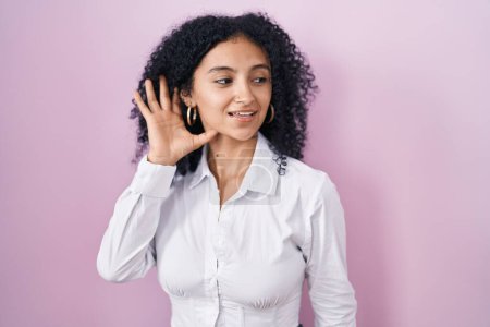 Téléchargez les photos : Hispanic woman with curly hair standing over pink background smiling with hand over ear listening an hearing to rumor or gossip. deafness concept. - en image libre de droit